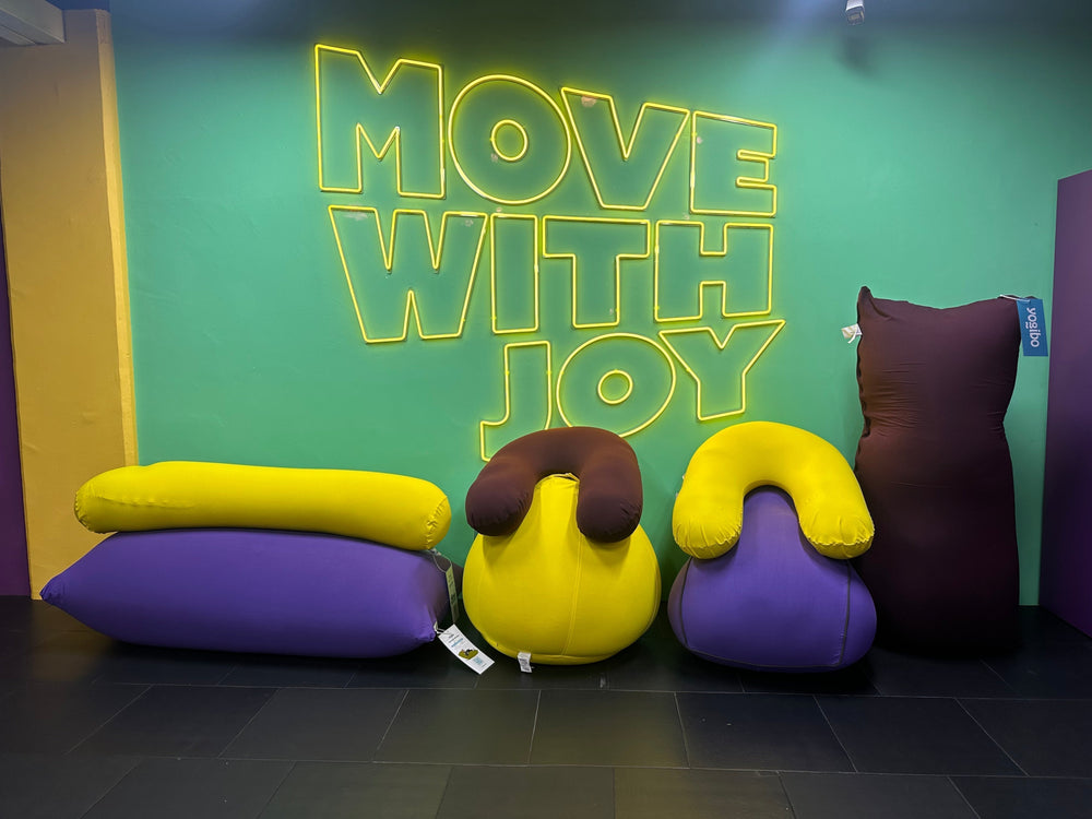 Collection of purple and yellow Yogibo bean bags arranged underneath a green neon Move With Joy sign