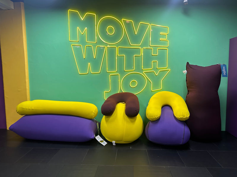 Collection of purple and yellow Yogibo bean bags arranged underneath a green neon Move With Joy sign